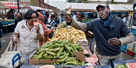Marktkoopman op Bijlmermarkt houdt stuk watermeloen omhoog, klant zet haar tanden in een stuk